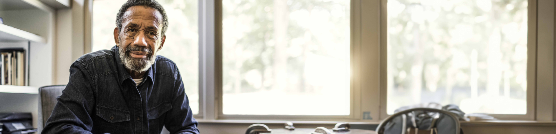 A man is sitting in front of bright windows and a book case.