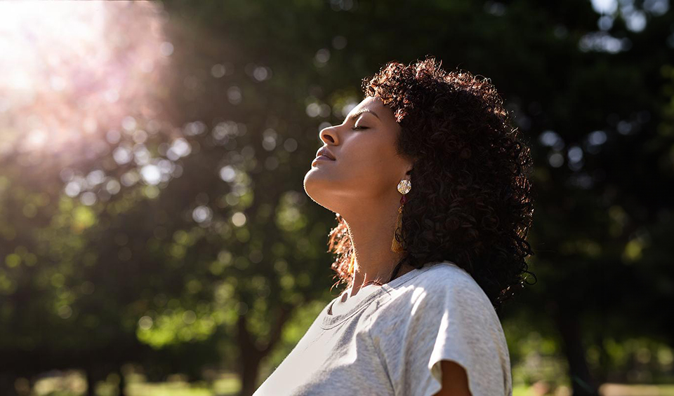 A woman stands with her head tilted back, being mindful and admiring the sun shining through the trees above her.