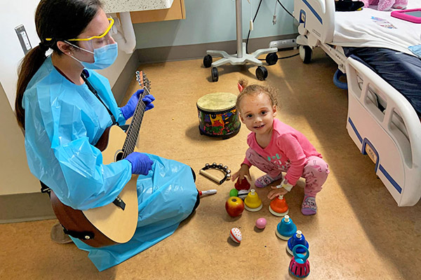 A young patient enjoys a music therapy session, playing with bells while a caregiver plays guitar