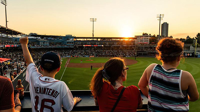 Young, cheering fans are sitting on the 1st base line of the Polar Park baseball field in Worcester at sunset.