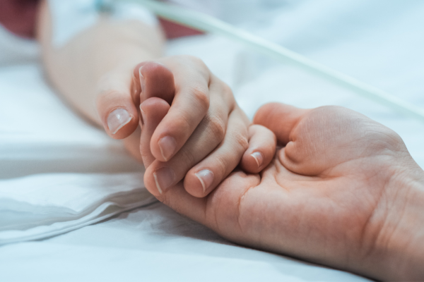 Close-up photo of someone holding a patient's hand. In the background, there is an IV in the patient's extended arm.