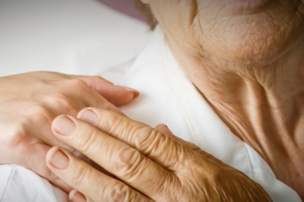 ﻿Close-up photo that represents the caring relationship between a health care worker and female hospice patient whose shoulder and jaw are the subject of the photo, along with the hand of the caregiver, positioned on the patient's shoulder, comforting her. The patient's hand is on top of the the caregiver's, in a way that shows appreciation for the comforting presence of her caregiver.