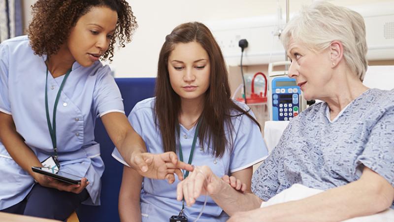 Two patient care associates speak with a woman in a hospital bed, highlighting their role in healthcare settings.