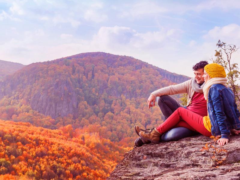 A couple enjoys the autumn view from a mountain peak, surrounded by vibrant fall foliage.