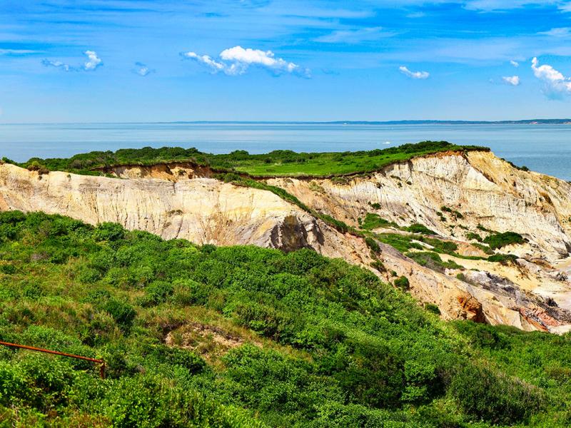 Photo of the New England Coast shows a large, green field, a lighthouse in the distance, and sandy cliffs that meet the ocean