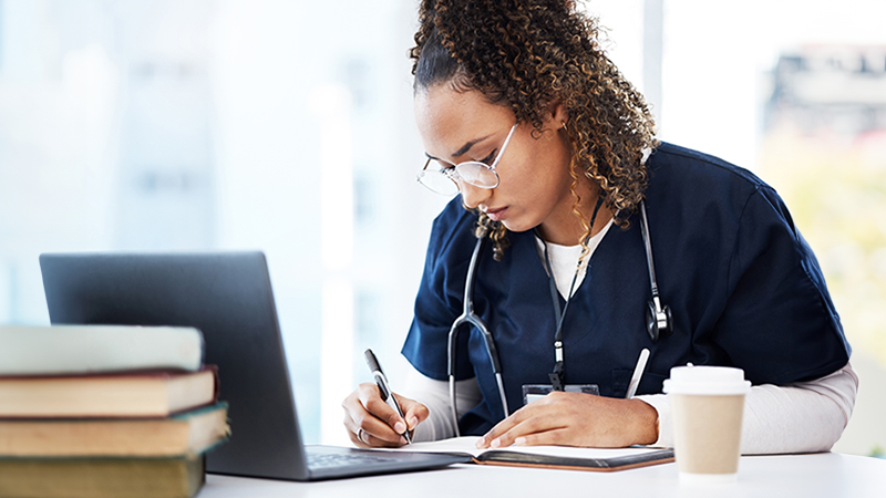 A radiologic technologist student takes notes in front of her laptop by a window inside a healthcare setting.