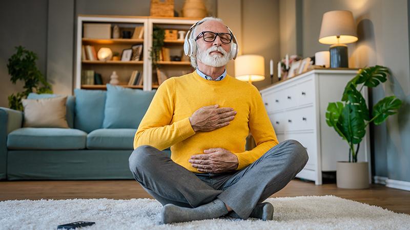A man sits cross-legged on a rug in his living room with his eyes closed, hands on his chest and abdomen, and headphone over his ears.