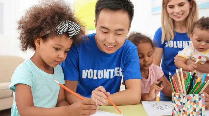 Young man volunteering at a daycare helping a young girl draw.