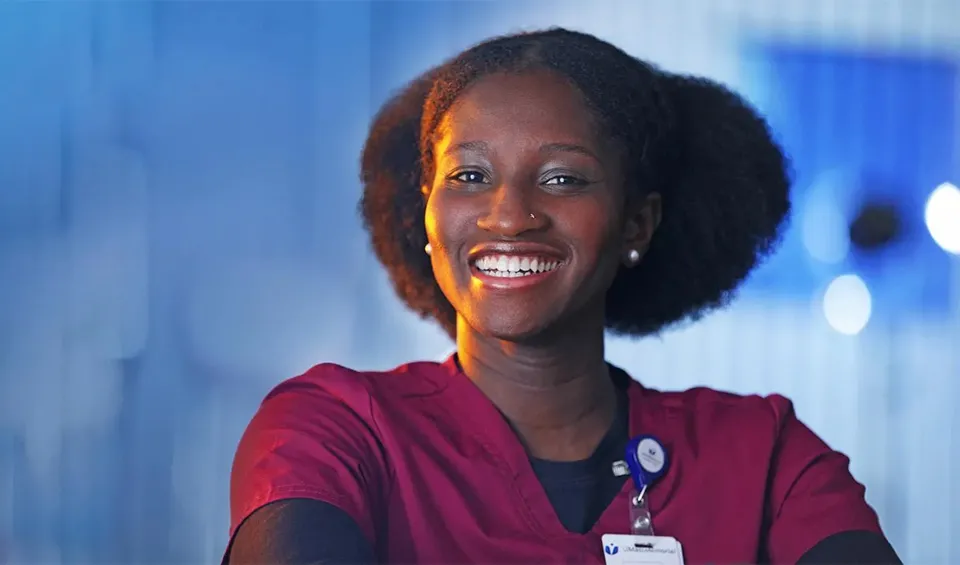 A smiling nurse in burgundy scrubs is smiling, on a blue background.