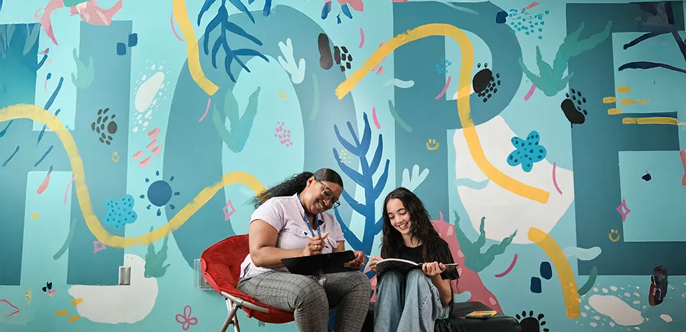 A woman with a clipboard is talking to a teenager with a notebook in front of a colorful floral wall.