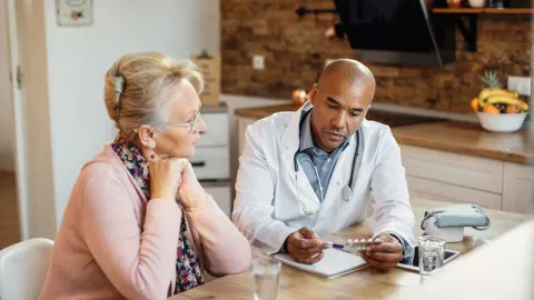 A doctor is explaining medication and care to a woman inside her kitchen.