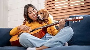 Woman sits on couch playing guitar while snuggling her dog