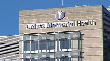 A bright blue sky shows behind the top of one of the health system's hospitals shows the UMass Memorial Health logo.