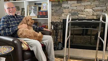 Marc Durocher recovering from hip surgery at home, in his comfortable chair, walker within reach, and his dog on his lap.