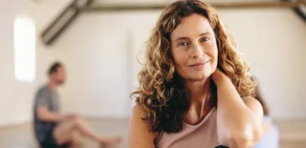 A woman with long hair smiles while sitting on the floor of a yoga, one knee to her chest, one hand behind her neck.
